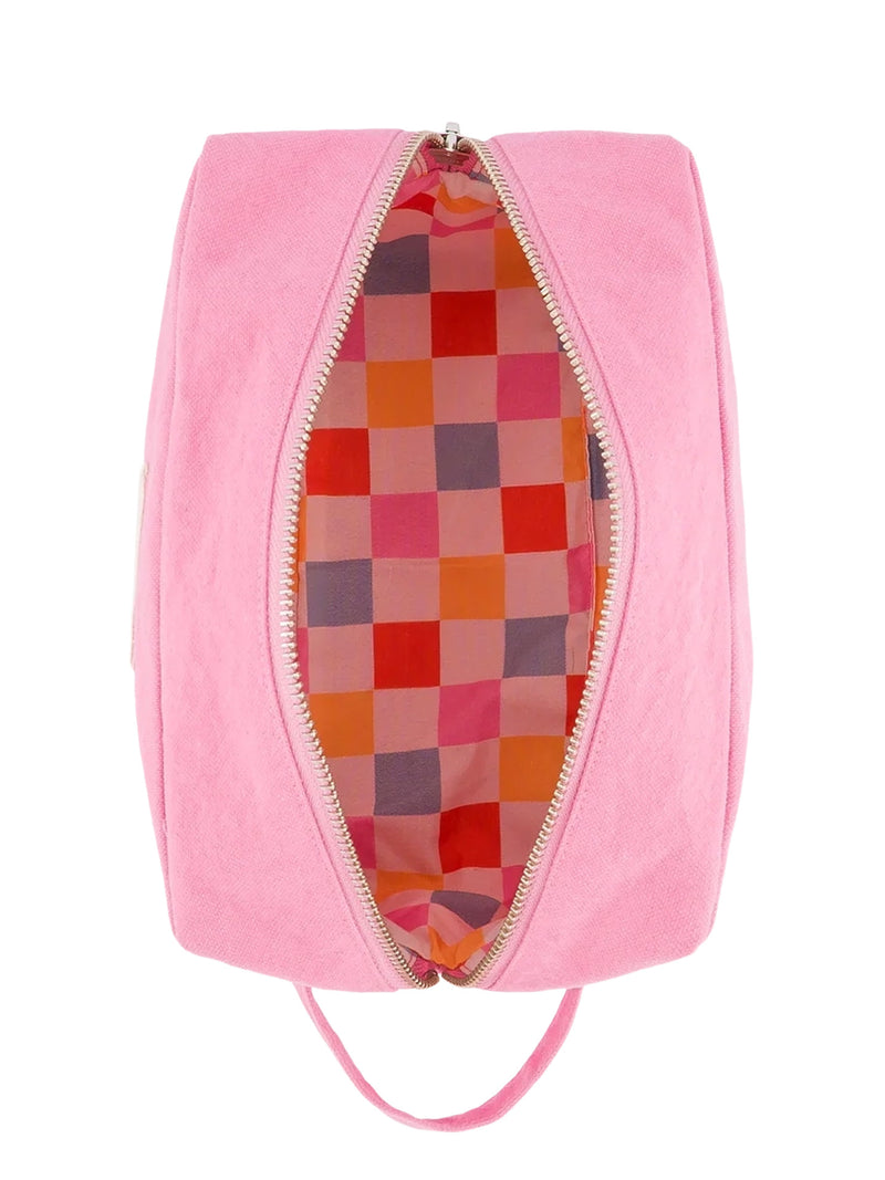 A pink zippered pouch with a generous hanging or carrying loop from the same pink canvas, shown open from the top, revealing a colorful checkered interior lining fabric in chess checkered pattern of red-orange, pink, light blue and off-white. The exterior material appears soft and slightly textured cotton canvas and the long zipper has silvery metal teeth.