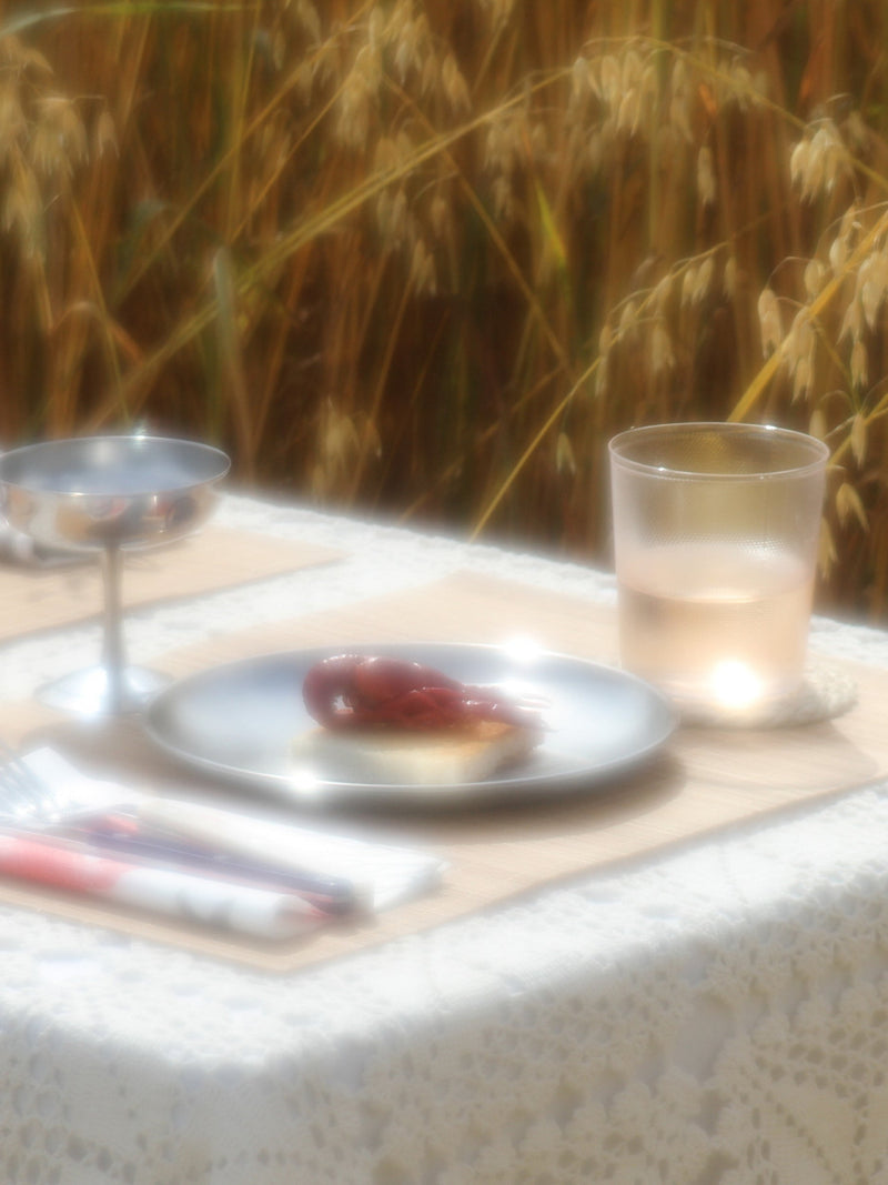 A softly blurred outdoor table setting with a white lace tablecloth, a plate holding a small red fruit and pastry, a glass cup, and a stemmed glass, with a warm field of tall grass in the background.