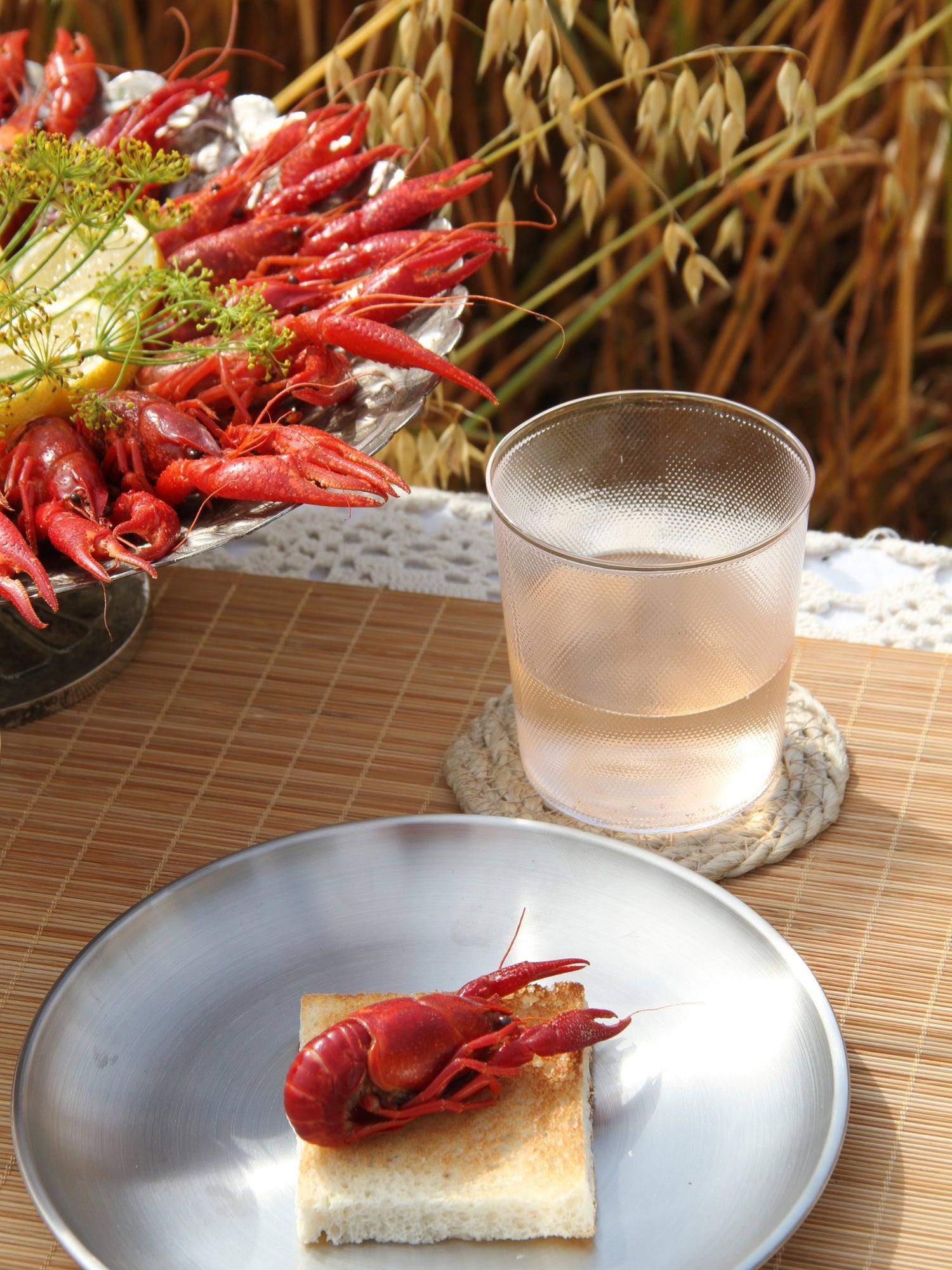A plated cooked crayfish on a piece of toast in the foreground, with a glass of pale beverage on a coaster and a tray of more crayfish in the background, set on a woven mat with a field of wheat behind it.