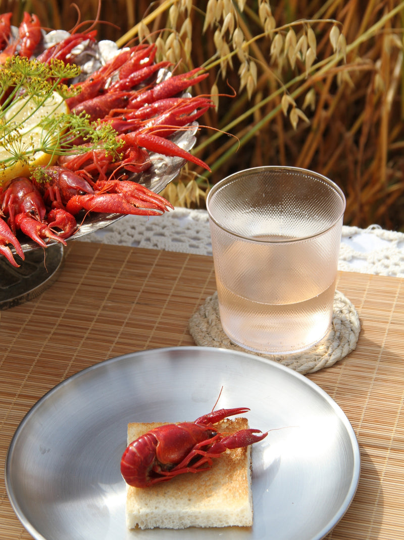 A plated cooked crayfish on a piece of toast in the foreground, with a glass of pale beverage on a coaster and a tray of more crayfish in the background, set on a woven mat with a field of wheat behind it.