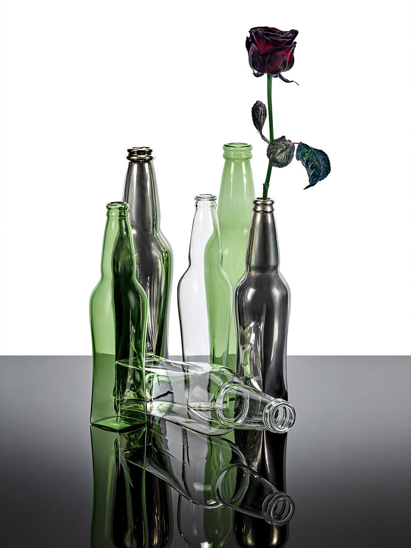 Collection of Josephine glass bottle vases in tall and small sizes with clear colour, green and metal chrome with a single red rose on a reflective table surface.