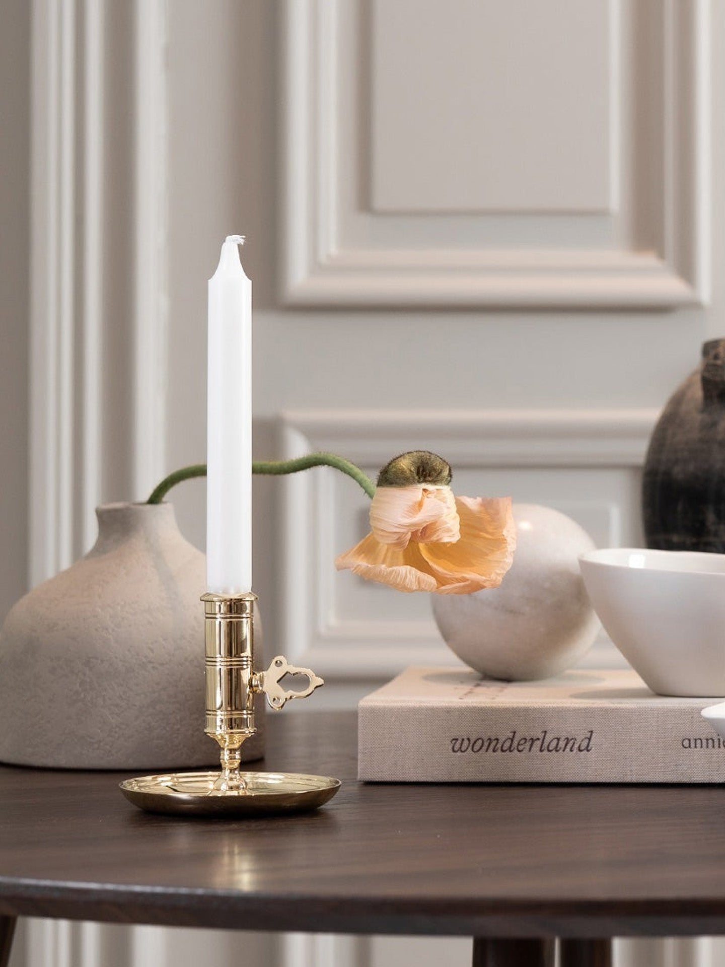 Decorative candle holder with a white candle on a table with books and vases in the background.