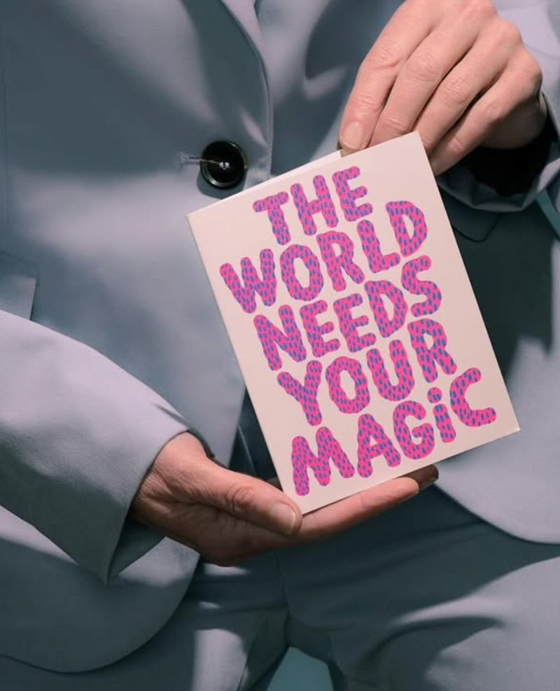 Person holding a card with 'The World Needs Your Magic' text in pink letters.