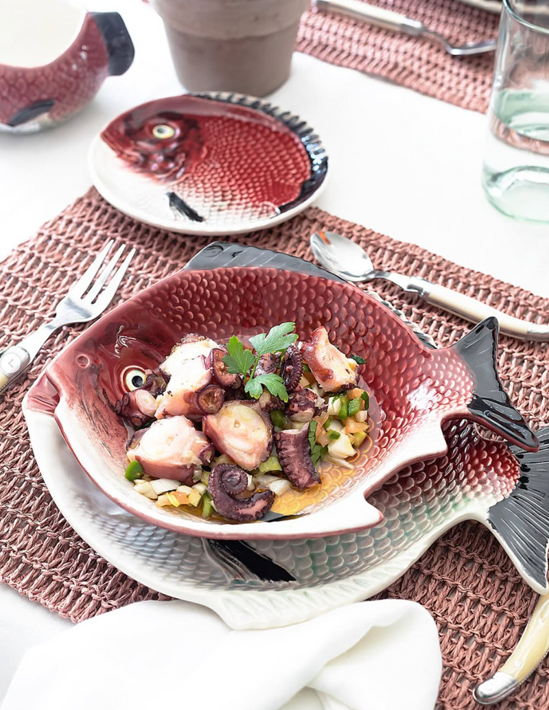 A ceramic fish-shaped bowl filled with octopus salad, placed on a table setting with a matching fish-patterned plate, woven placemats, silverware, and a glass.