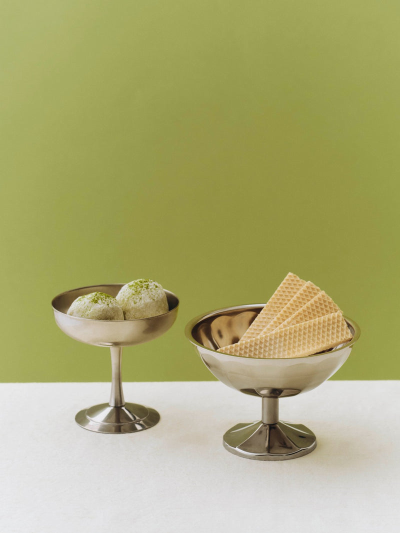 Two metal bowls with ice cream and wafers on a white surface against a green background