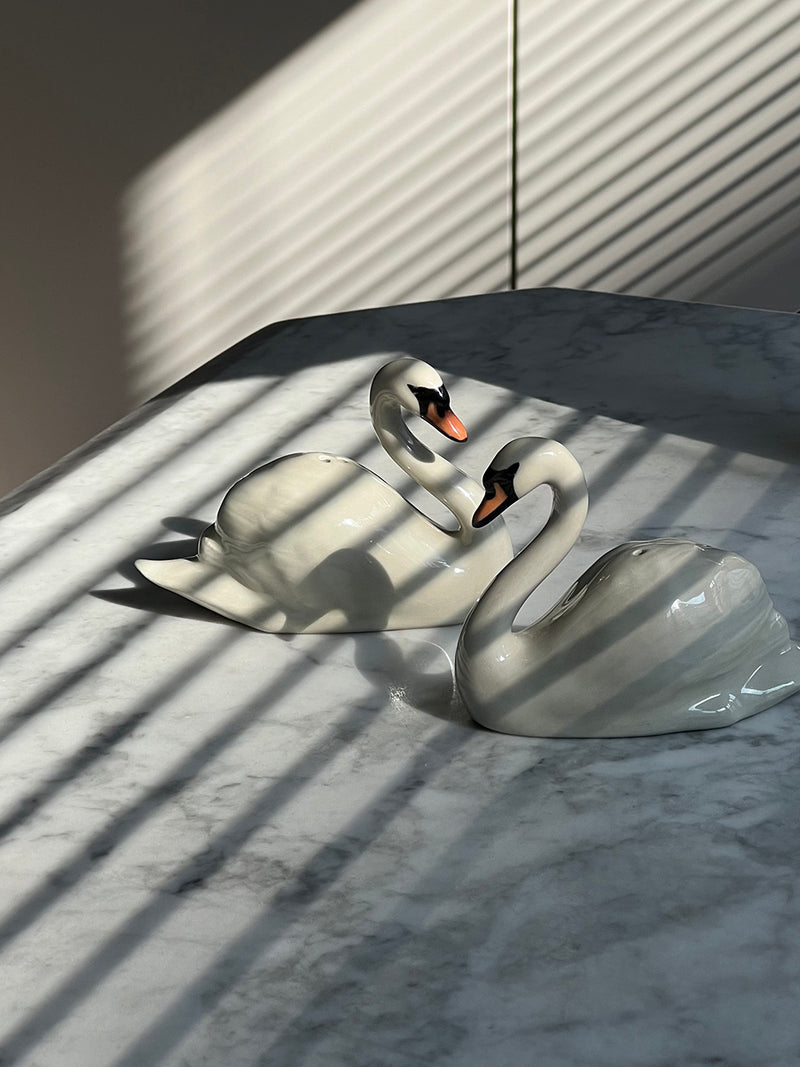 Two white swan shakers with orange beaks resting on a gray marble surface under diagonal window blinds casting shadows.