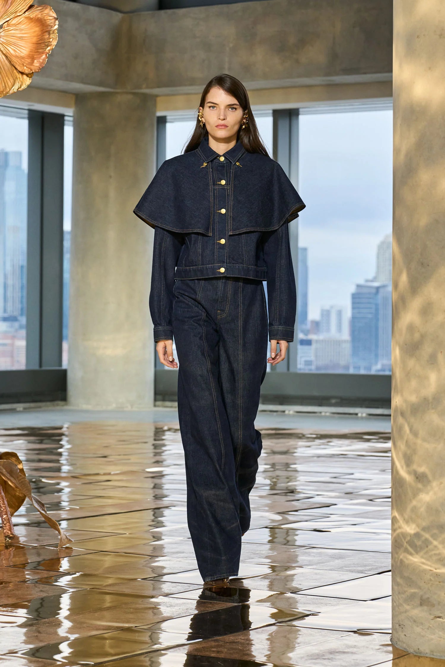 Model wearing a dark denim outfit on a runway with a cityscape background
