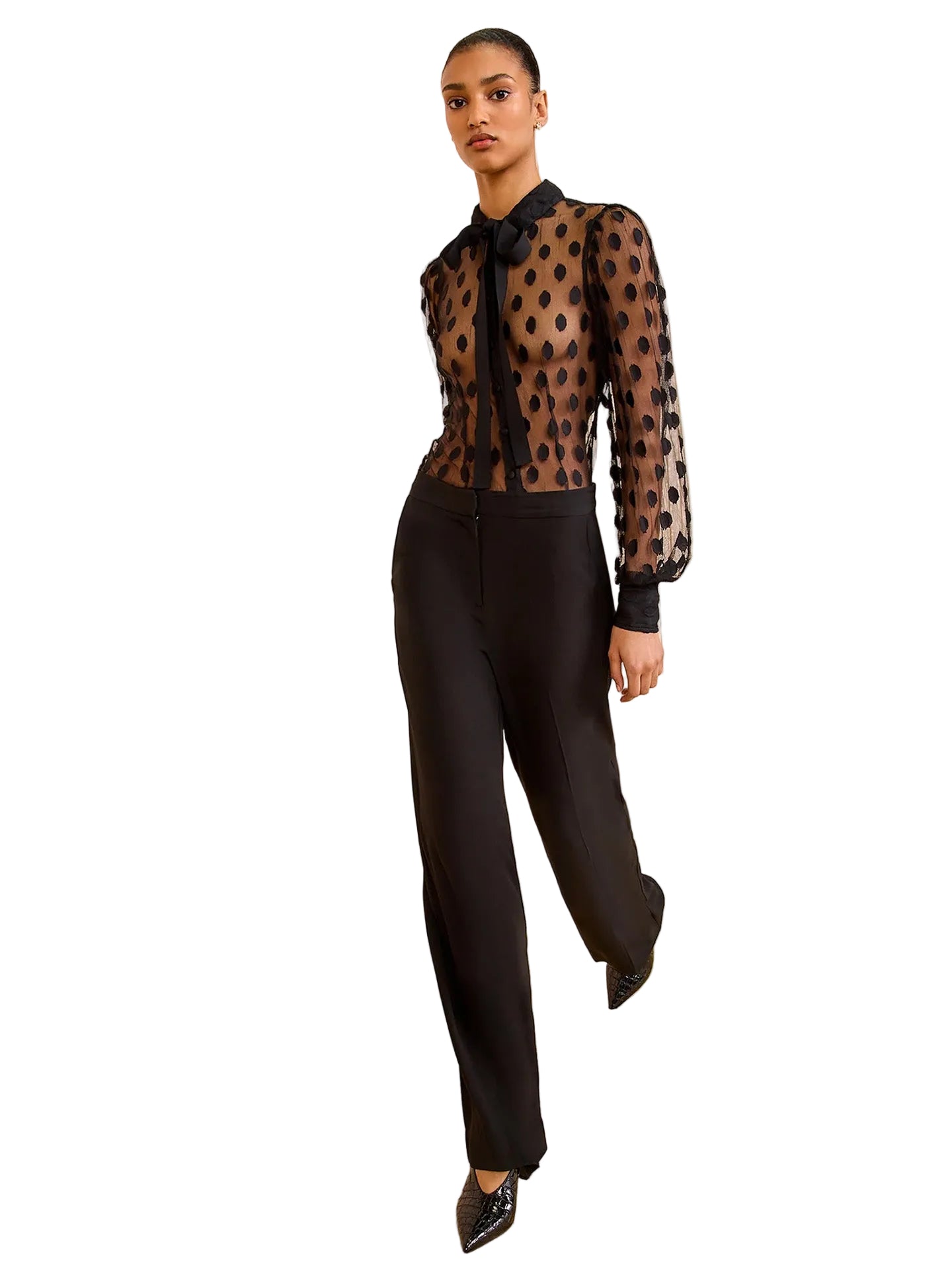 Woman wearing a sheer black polka dot blouse with black pants against a beige wall.