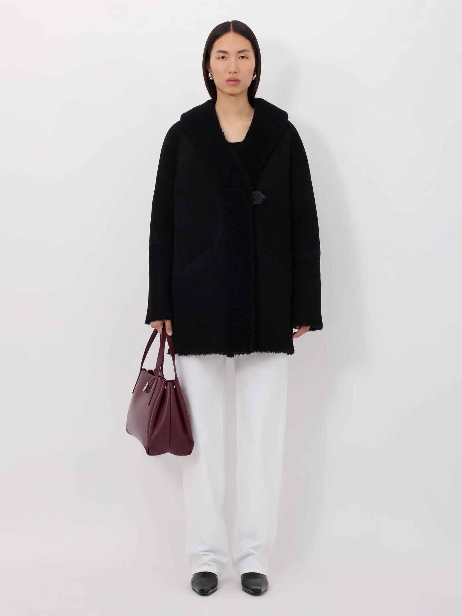 Person wearing a black shearling coat and white pants holding a maroon handbag on a white background