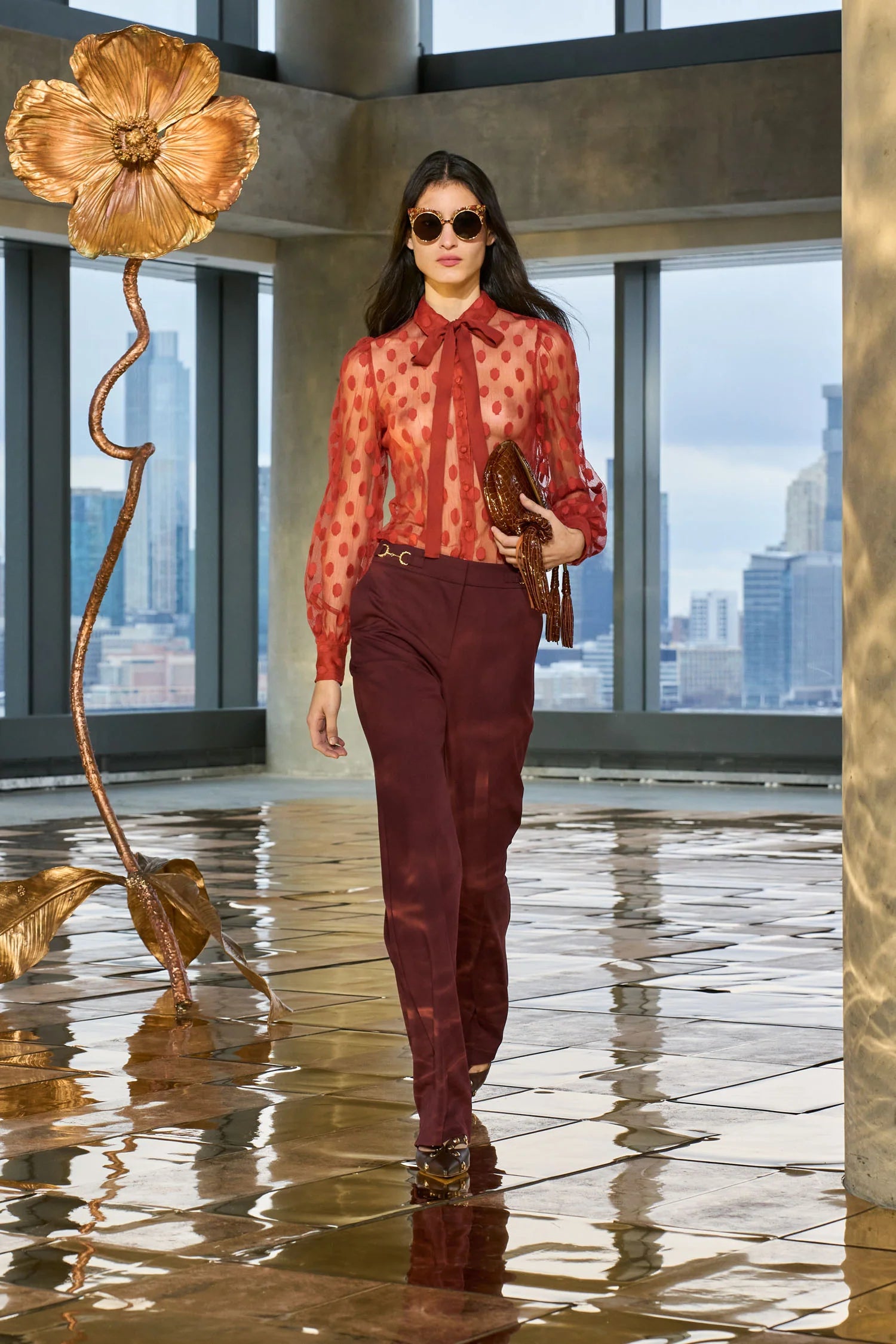 Woman in a red blouse and burgundy pants walking in a modern indoor setting with large windows and artistic sculptures.