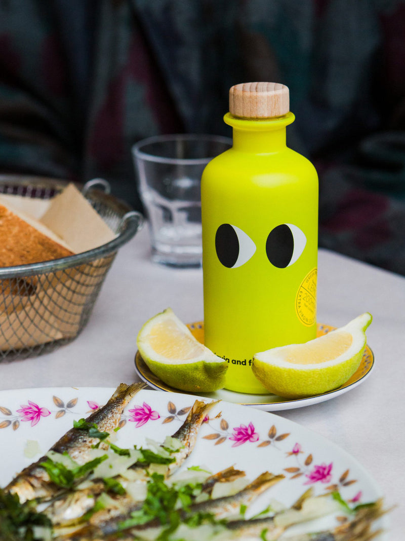 Green bottle with sideways looking eye design on a table with lemon slices and friend little fish, bread and water glass on a table.