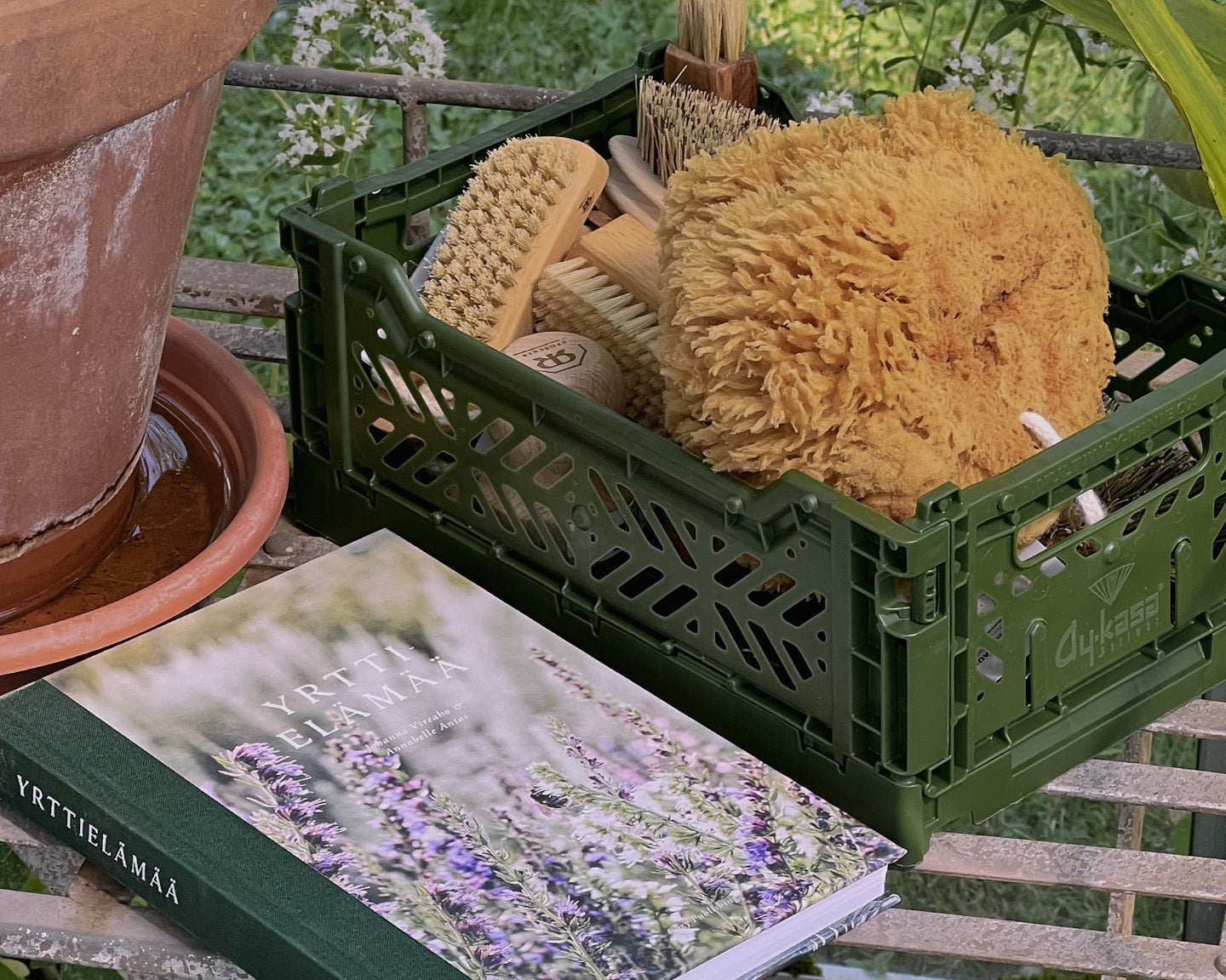 XS sized Aykasa storage crate in moss green filled with Bürstenhaus Redecker's nailbrushes, sponges and other cleaning brushes, next to a book named Yrttielämää and a ceramic plant pot on metal table or chair