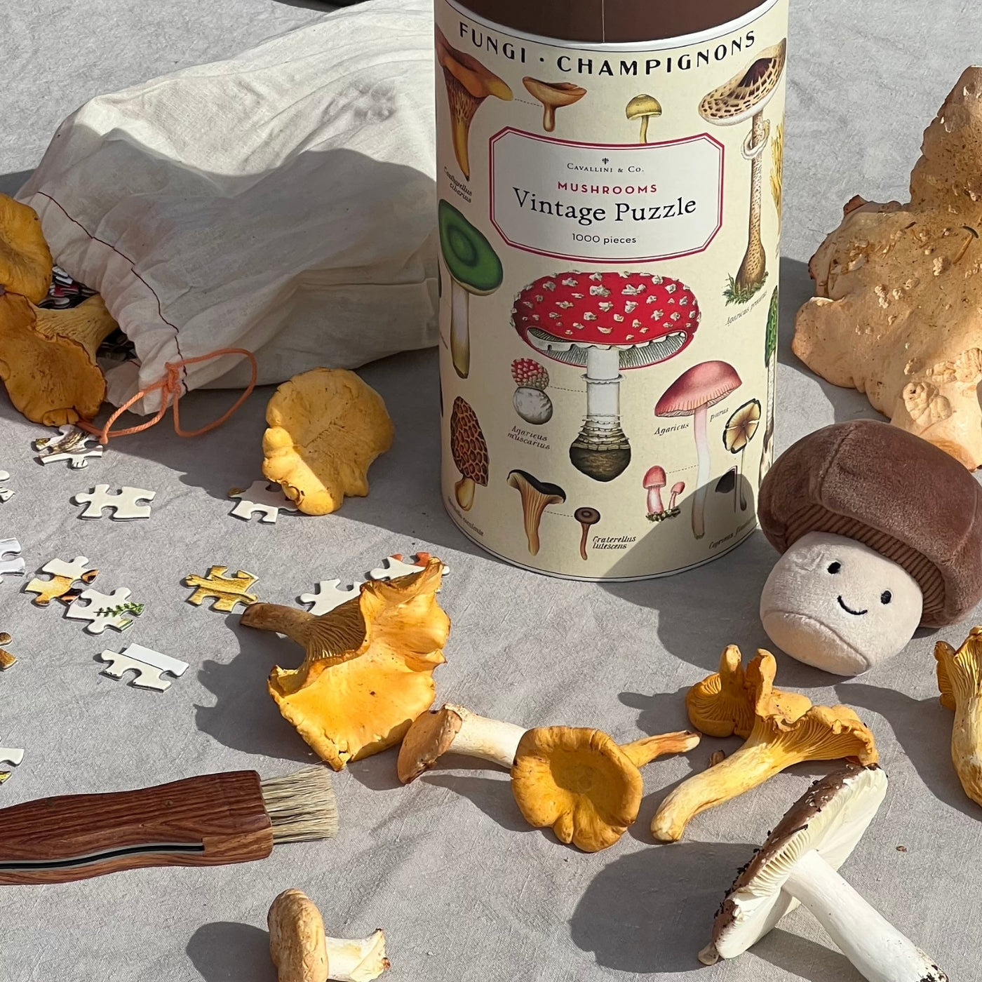 A vintage-style mushroom-themed jigsaw puzzle box labeled 'Fungi - Champignons Cavallini & Co Mushrooms Vintage Puzzle 1000 pieces' stands on a grey fabric surface surrounded by loose puzzle pieces, yellow chantarelle mushrooms, a small brown plush Jellycat mushroom soft toy, a mushroom brush, and jigsaw puzzle piece fabric pouch.