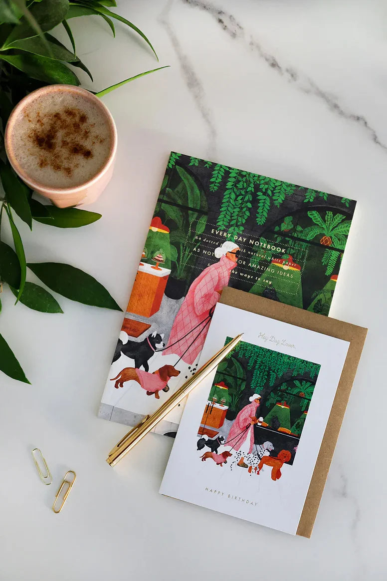 Two greeting cards with a woman and dogs design, a pen, and a cup of coffee on a marble surface.