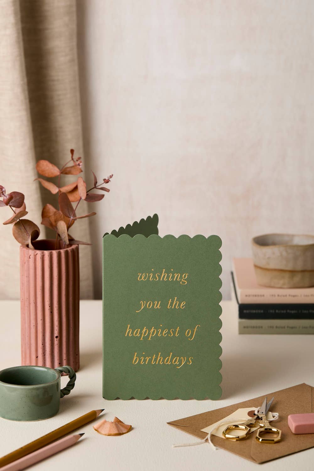 Green scalloped-cut birthday card with gold text 'wishing you the happiest of birthdays' standing ope on a desk with a vase, books, and stationery.