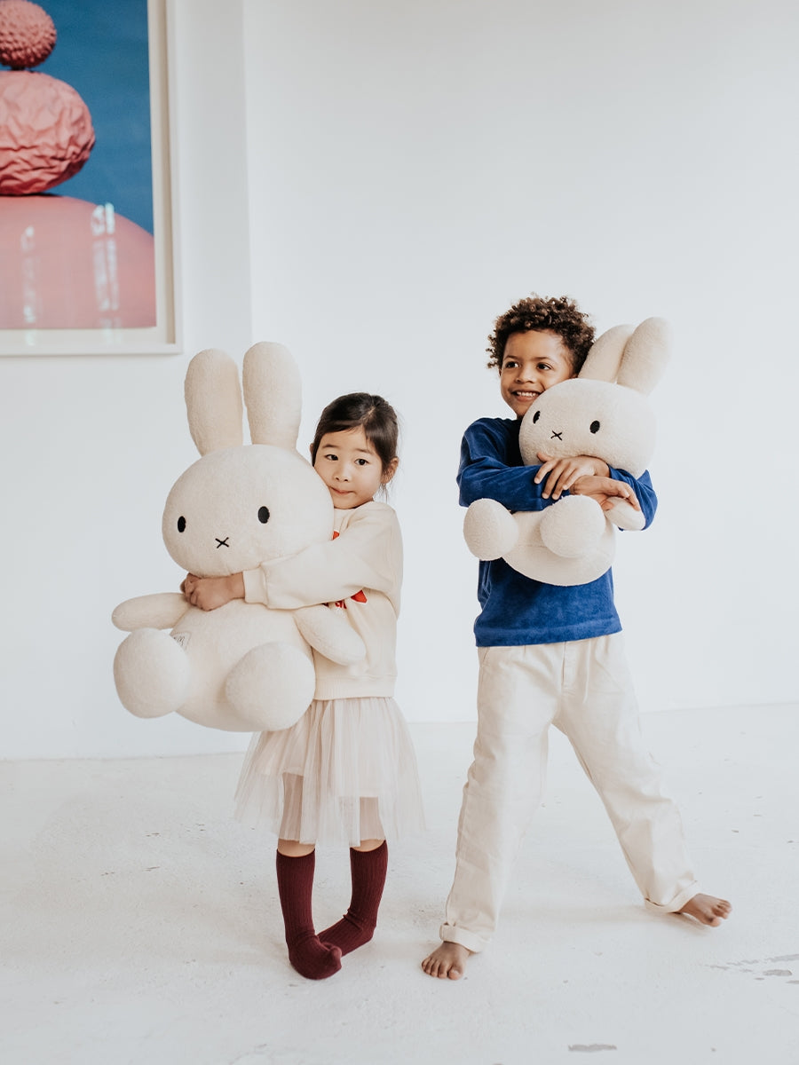 Two children holding large plush bunnies in a minimalistic room.