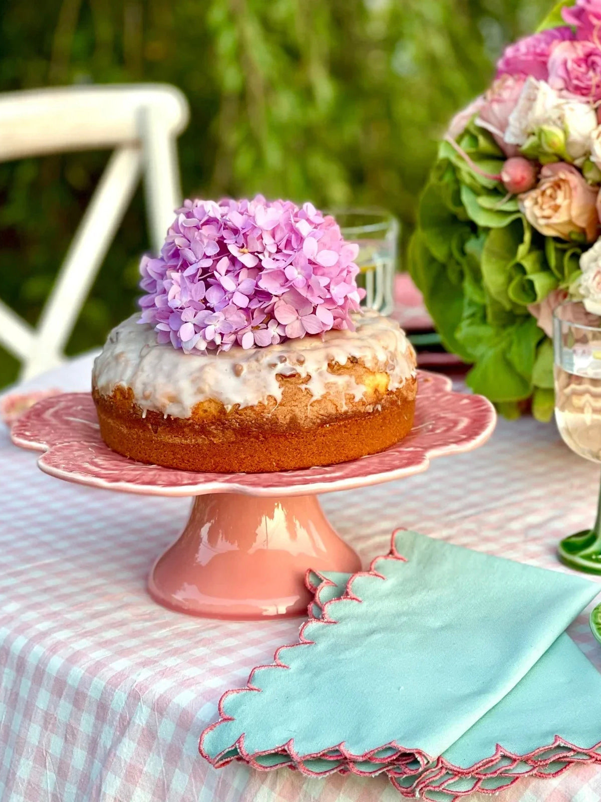 Cake on a pink stand with pink flowers on top, set on a table with a checkered tablecloth.