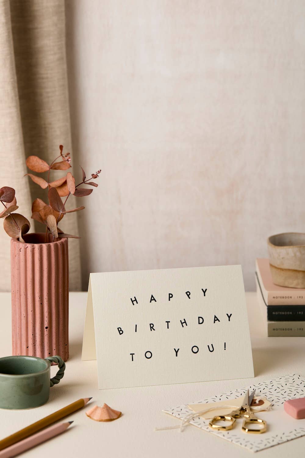 A white standing greeting card with black text reading 'HAPPY BIRTHDAY TO YOU!' on a tabletop, styled with a pink ribbed vase, dried leaves, pencils, books, and small accessories in a soft neutral room setting.