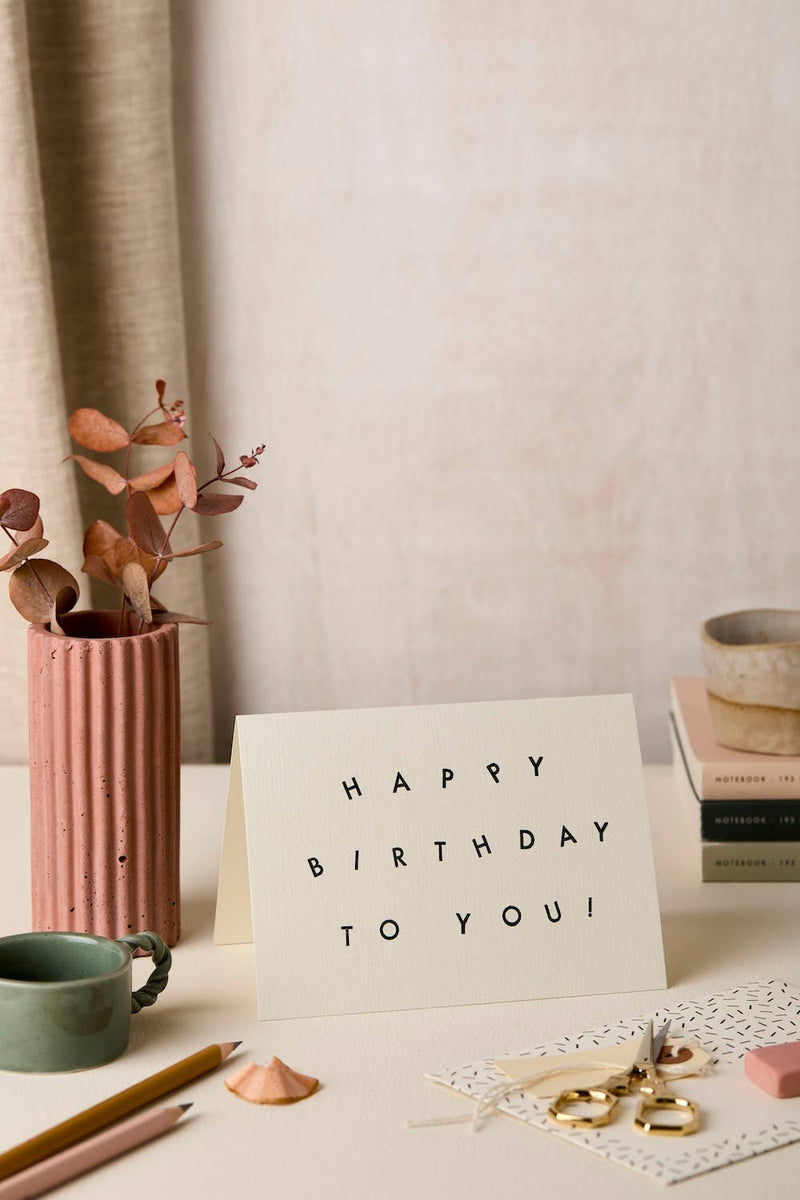 A white standing greeting card with black text reading 'HAPPY BIRTHDAY TO YOU!' on a tabletop, styled with a pink ribbed vase, dried leaves, pencils, books, and small accessories in a soft neutral room setting.