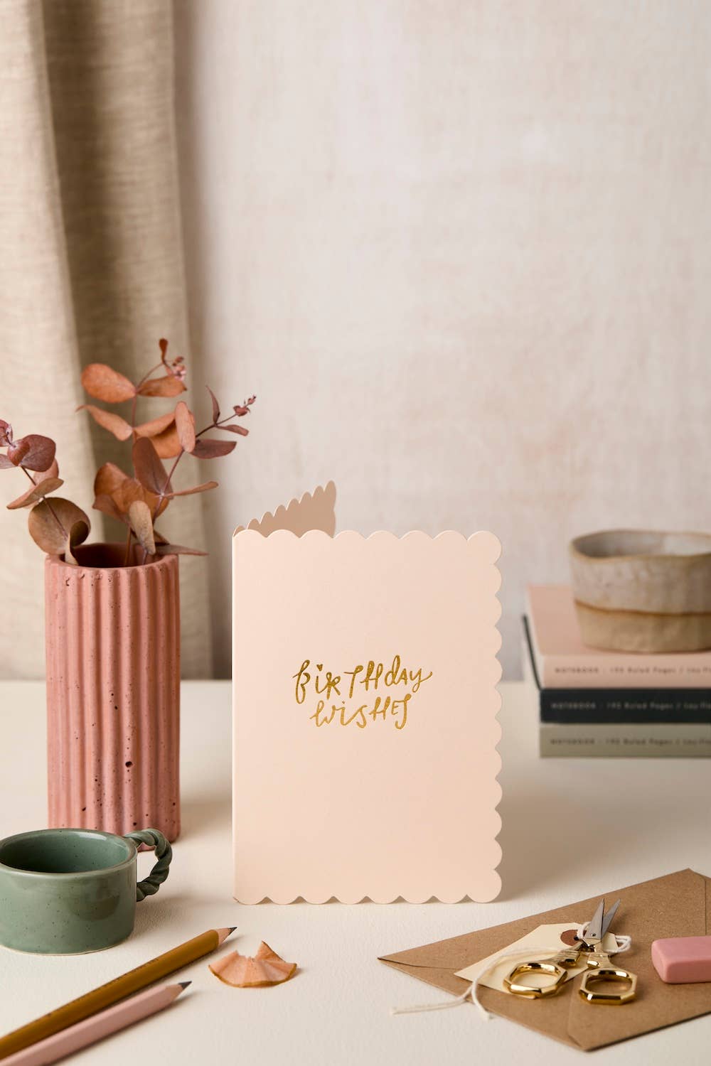 Soft pink birthday card with gold text 'Birthday wishes' on a table with a pink vase, green mug, and books.