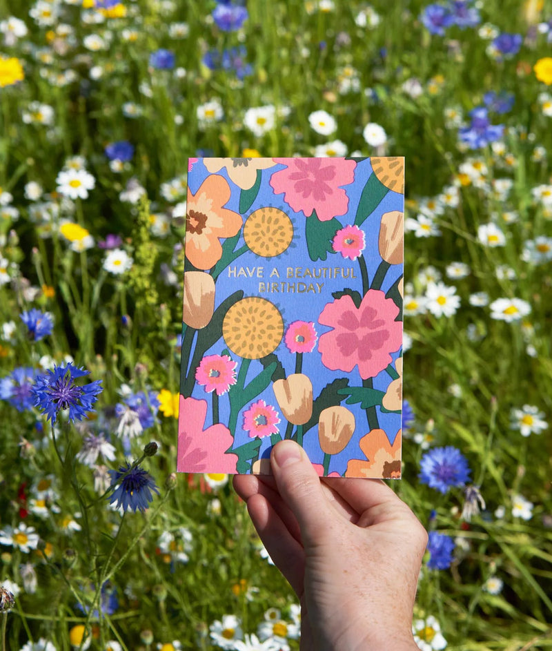 Hand holding a colorful floral birthday card in front of a field of wildflowers