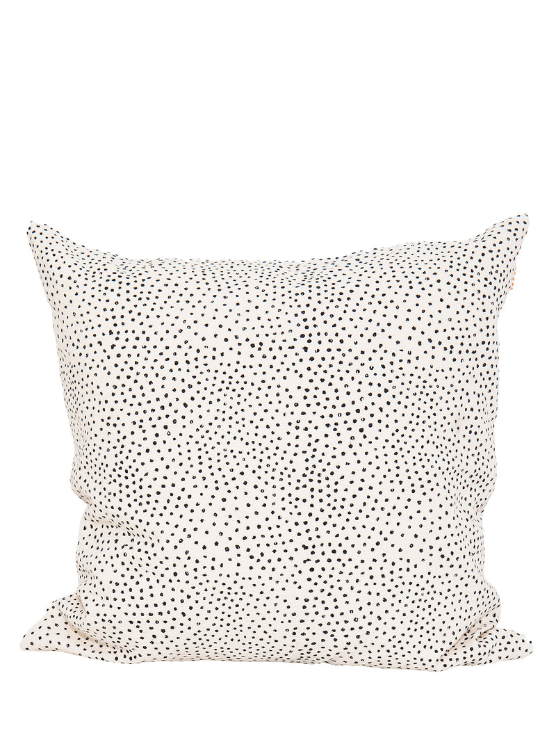 A cushion cover with a white background and black irregular dots pattern. The cover is displayed in a relaxed, casual setup.