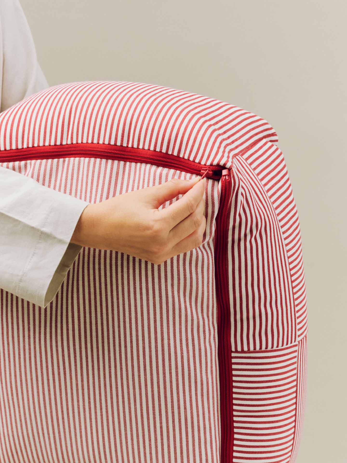 A close-up of a person's hand unzipping HAY's new dog bed with red and white vertical stripes and a red zipper at the bottom.