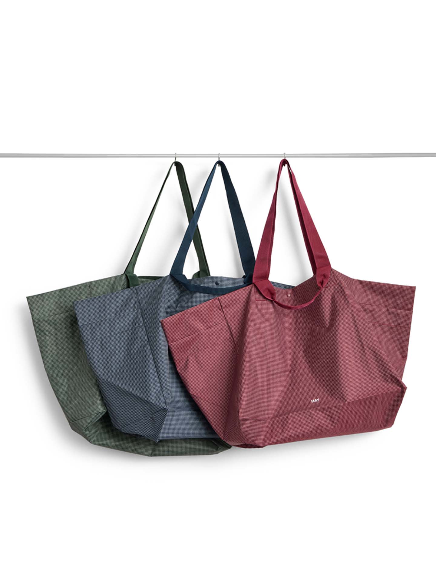 Three reusable tote bags hanging from a horizontal bar against a white background: an olive green bag at the back, a slate blue-gray bag in the middle, and a dusty rose red bag in the foreground.