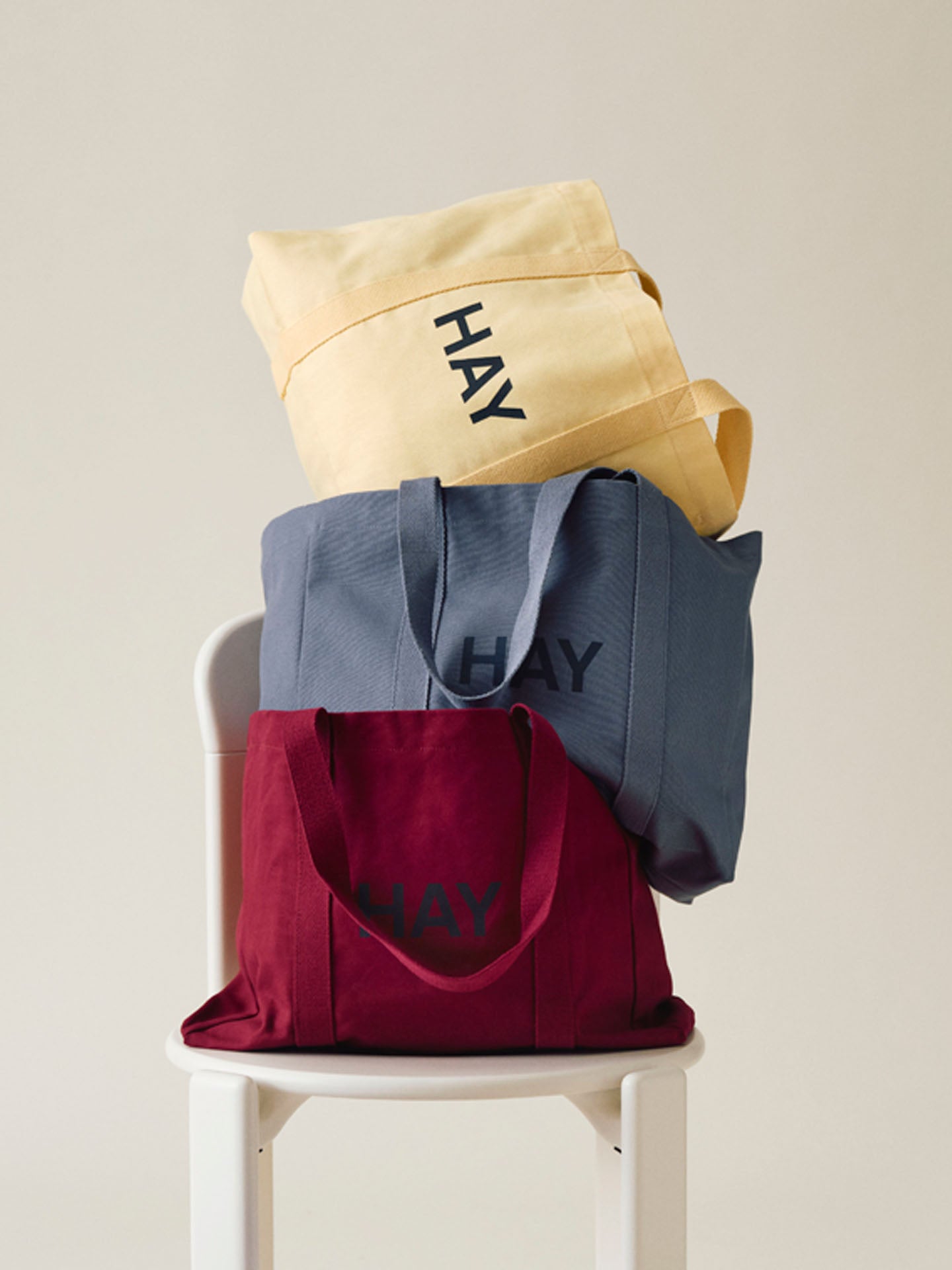 Three canvas tote bags stacked on a white chair against a light beige background. The solid coloured bags are in pale yellow, slate blue, and deep burgundy, each printed with the word 'HAY' in dark lettering.