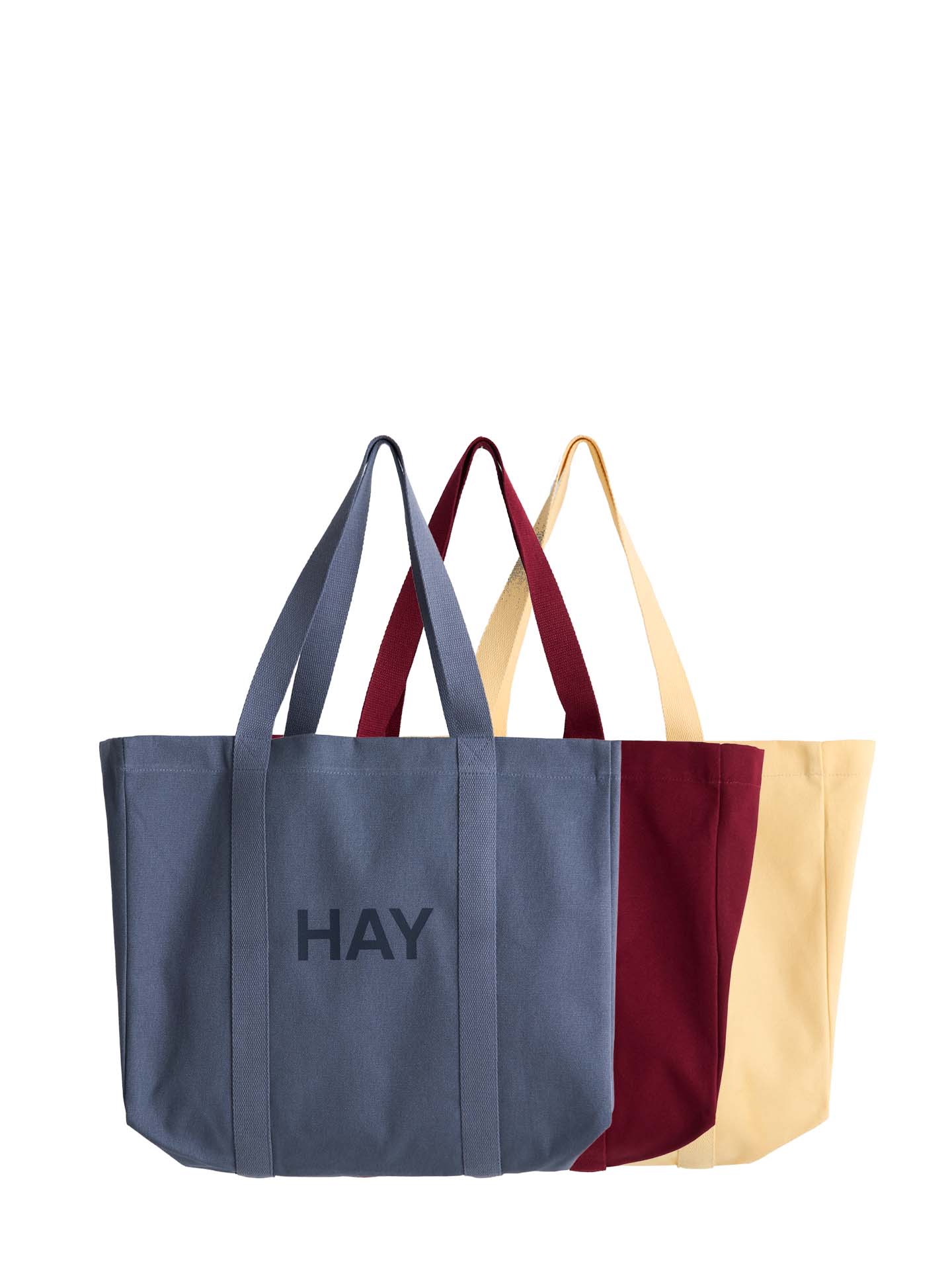 Three canvas tote bags in slate blue, burgundy, and cream, arranged in a row against a plain white background. The front slate blue bag features the word 'HAY' printed in dark gray on the front.