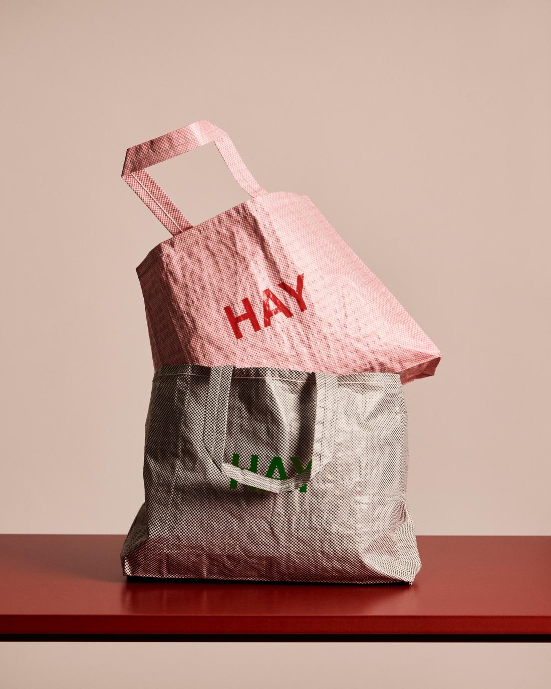 Two tote bags, one pink and one gray with 'HAY' branding, on a red surface.