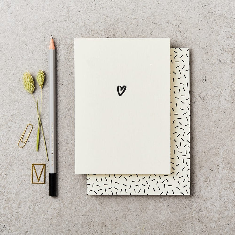 Love greeting card with a heart drawing, pencil, paper clips, and dried plants on a concrete surface