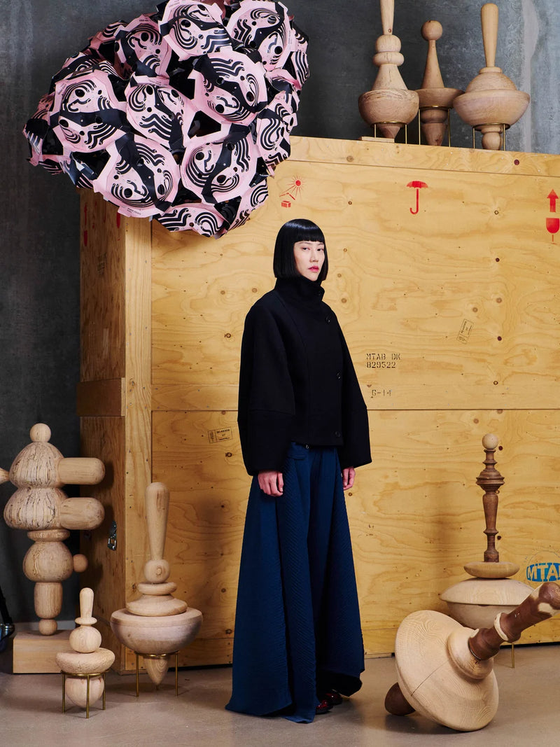 A model stands in a studio setting wearing a black hooded long-sleeve top and dark navy wide-leg trousers. The background features a large wooden crate with a pink and black patterned fabric draped over it, surrounded by various wooden sculpture props.
