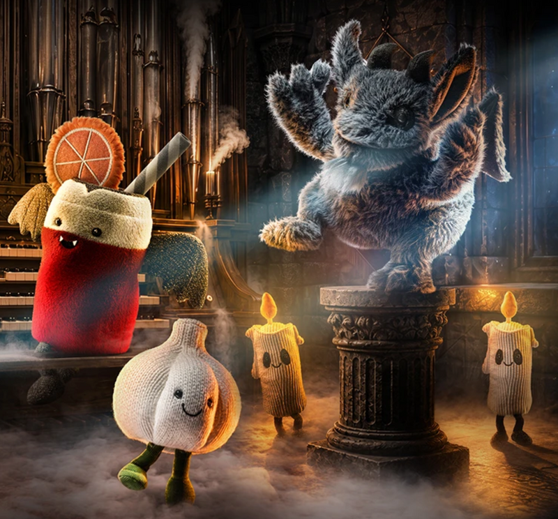 A whimsical illustrated scene showing a group of anthropomorphic fantasy characters in a dark gothic interior with candles and mist. The main subject is a large furry gray creature standing on a pedestal, with other characters including a red drink-like figure, a garlic character, and candle figures around it.