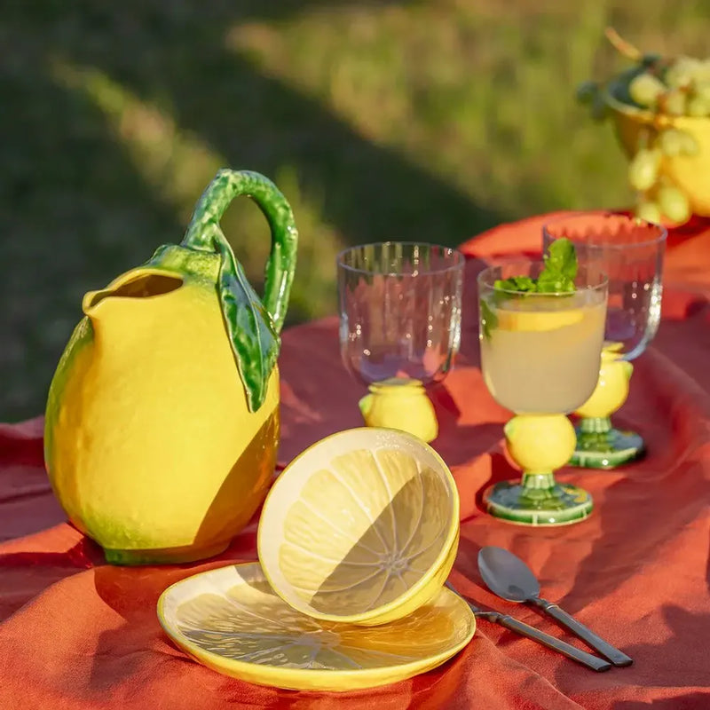Yellow ceramic lemon-shaped pitcher and glasses on a red tablecloth with a blurred green outdoor background.