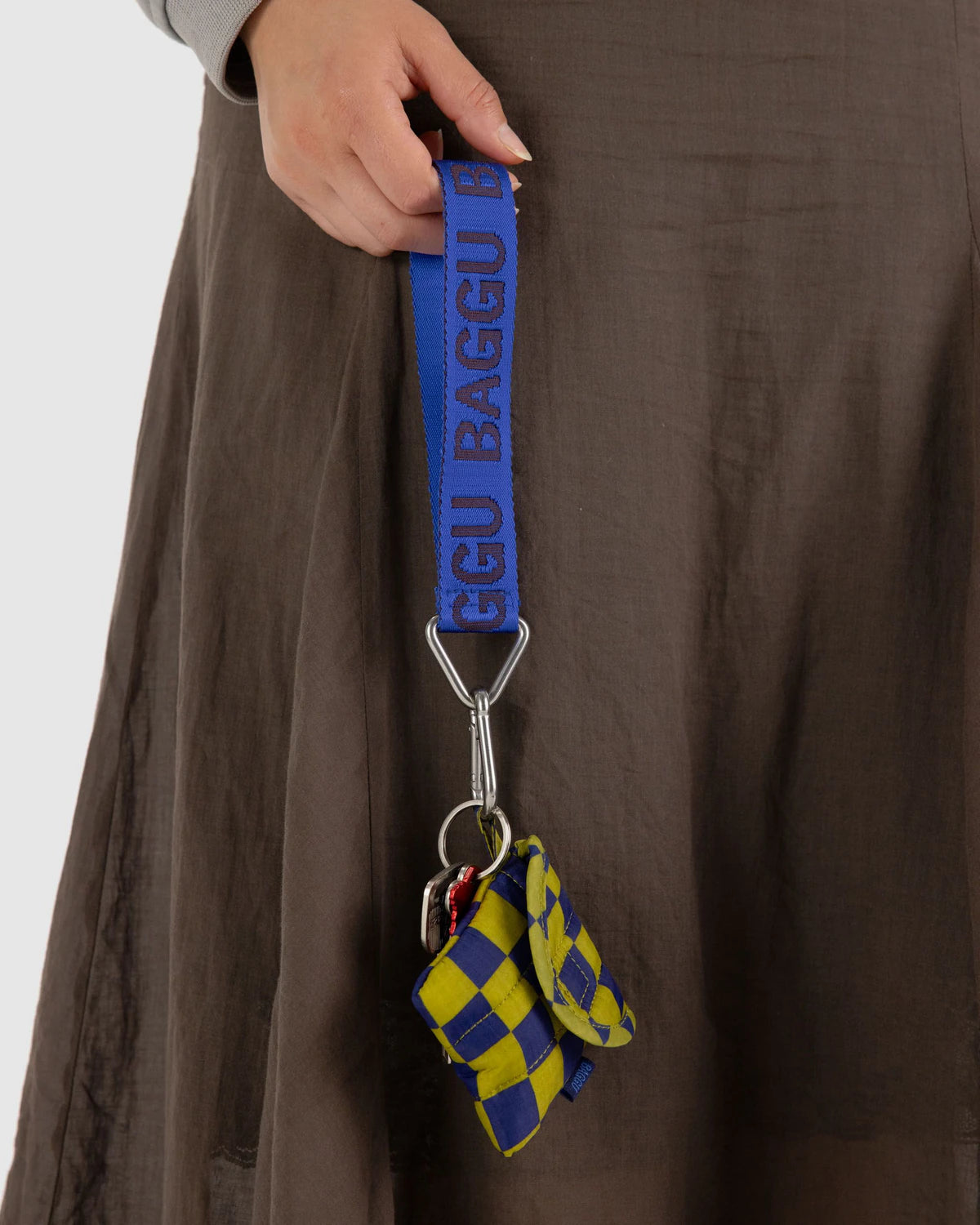 Hand holding a blue baggu strap with a small bag attached, against a brown background.