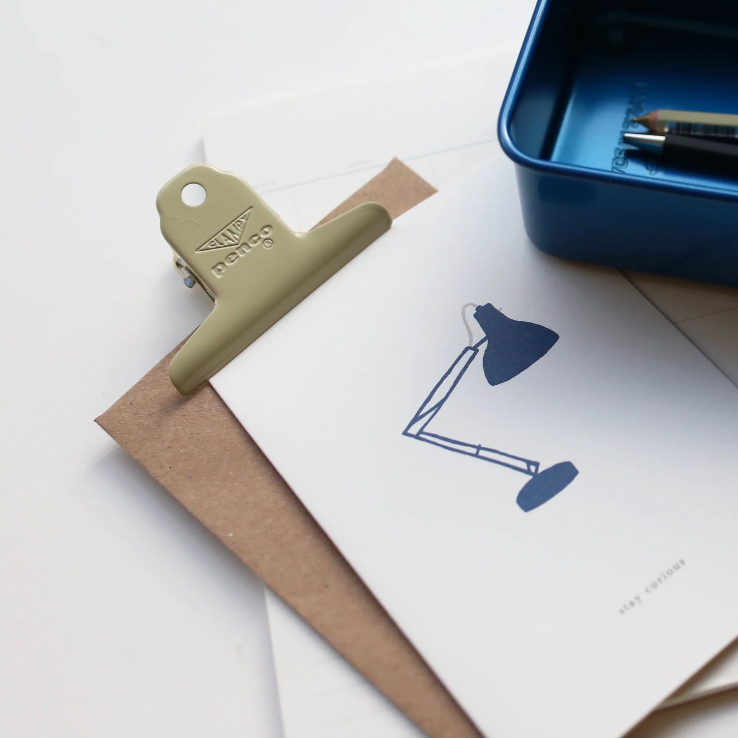 Green clip on a piece of paper with a blue lamp illustration, next to a blue metal tin.