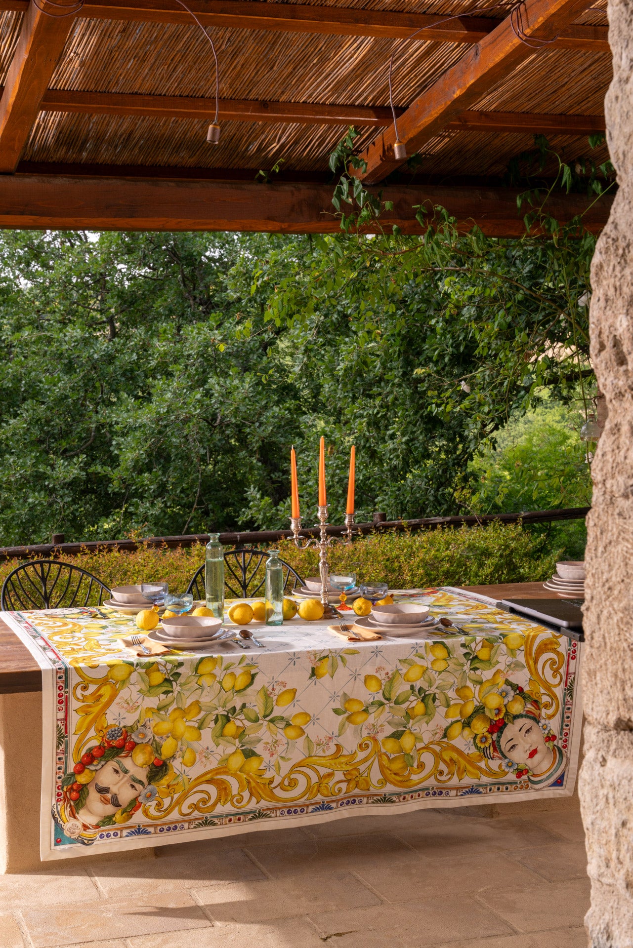 Outdoor dining table with decorative tablecloth and candles, surrounded by greenery.