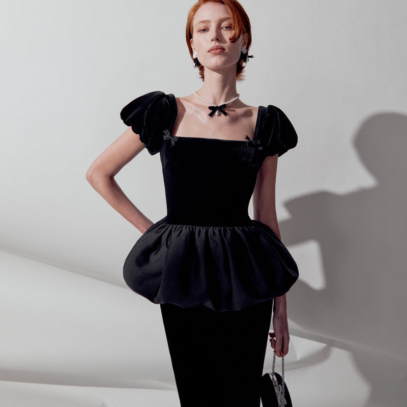 A model wearing a black velvet peplum top with puffed cap sleeves, a square neckline, and delicate black bow details on the shoulders, paired with black trousers and a black choker necklace.