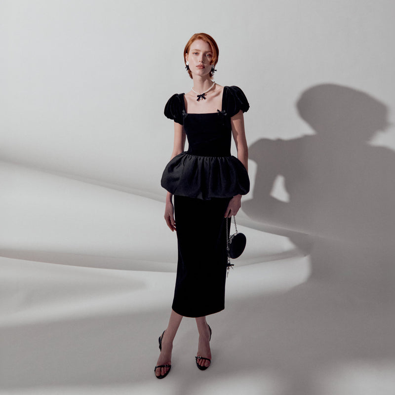 A model with red hair stands in a studio wearing a black velvet dress with puff sleeves, a square neckline, and a ruffled peplum waist. She is holding a small black handbag and wearing black strappy heels against a white background with a prominent shadow.