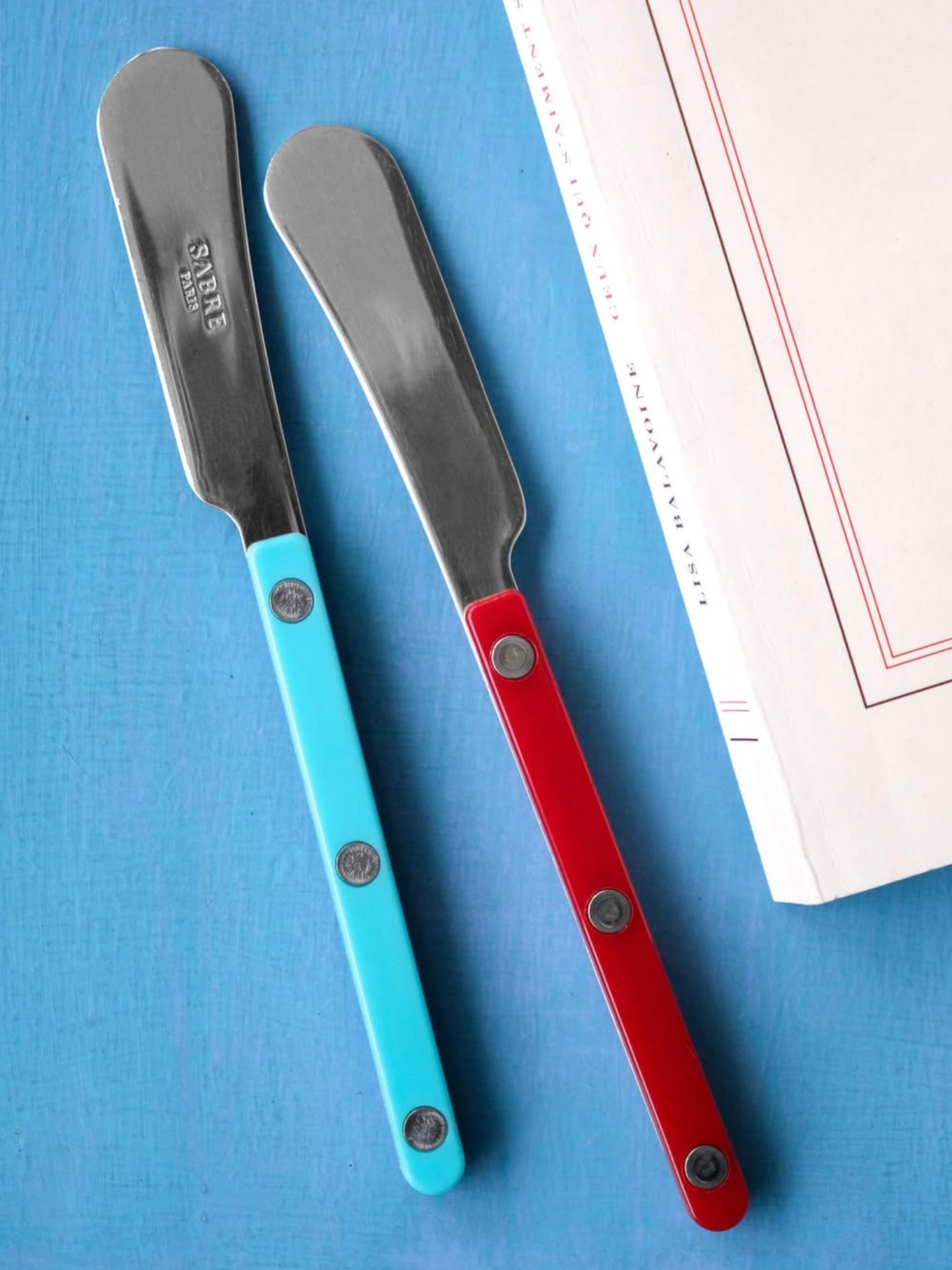 Two butter spreaders with blue and red handles on a blue surface.