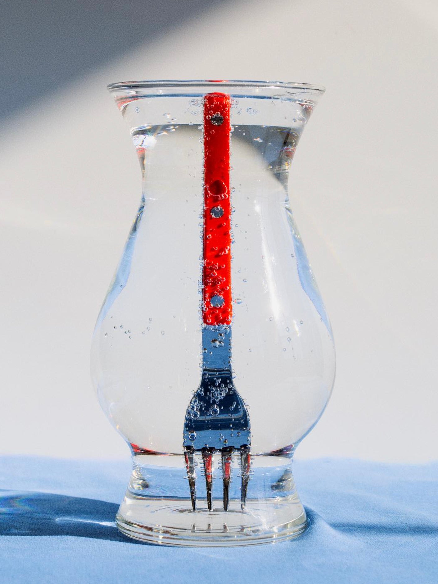 Red fork inside a clear glass vase on a blue surface with a white background