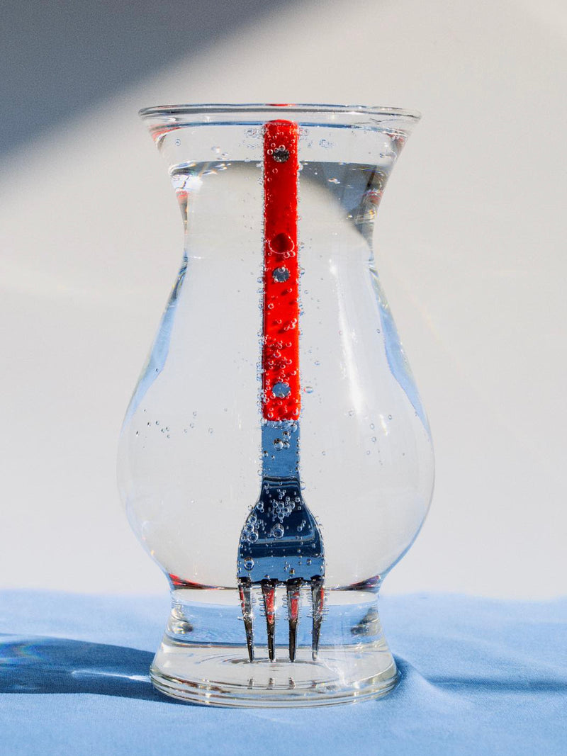 Red fork inside a clear glass vase on a blue surface with a white background