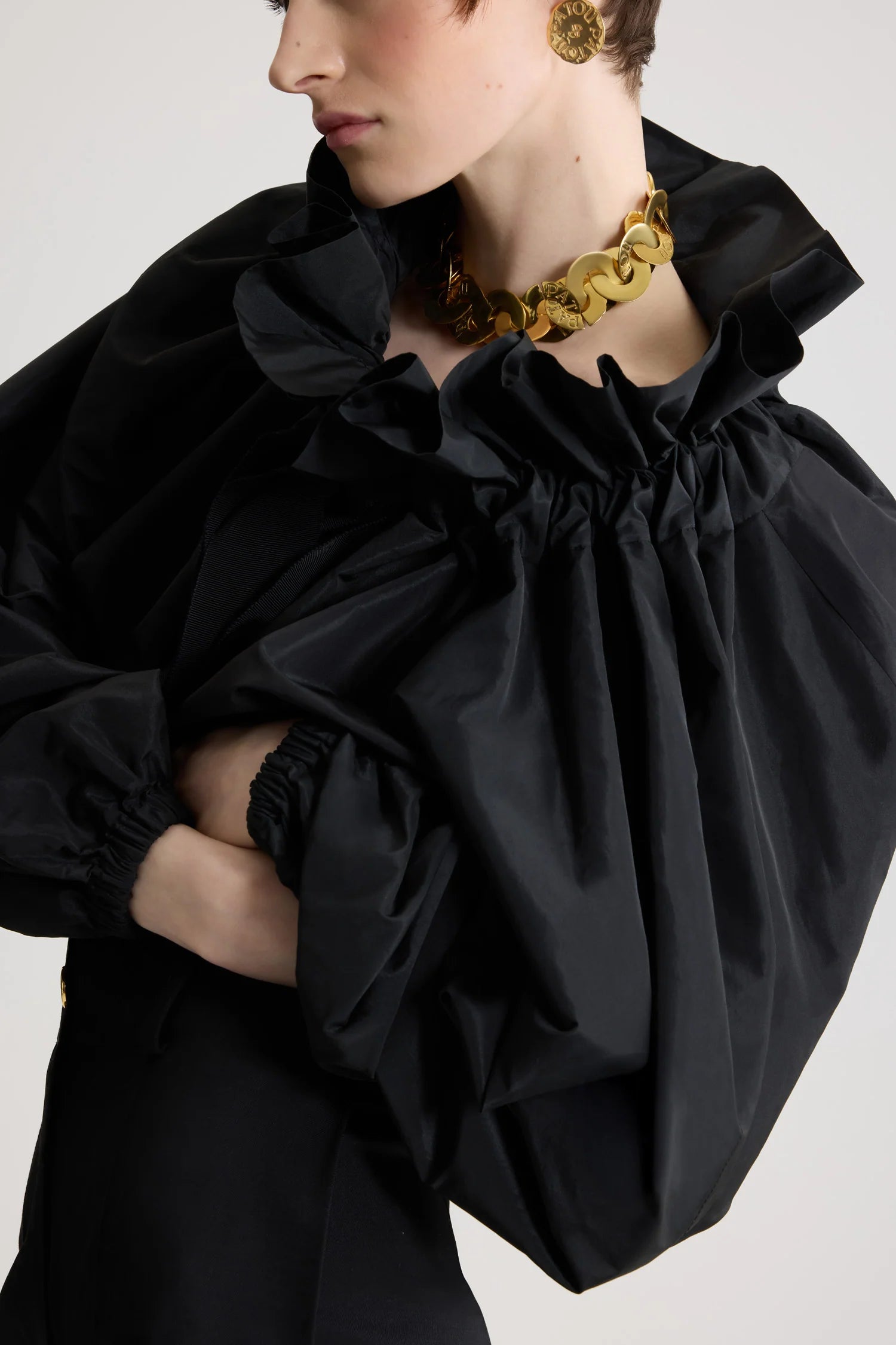 Person wearing a black outfit with ruffled sleeves and a gold necklace on a light gray background