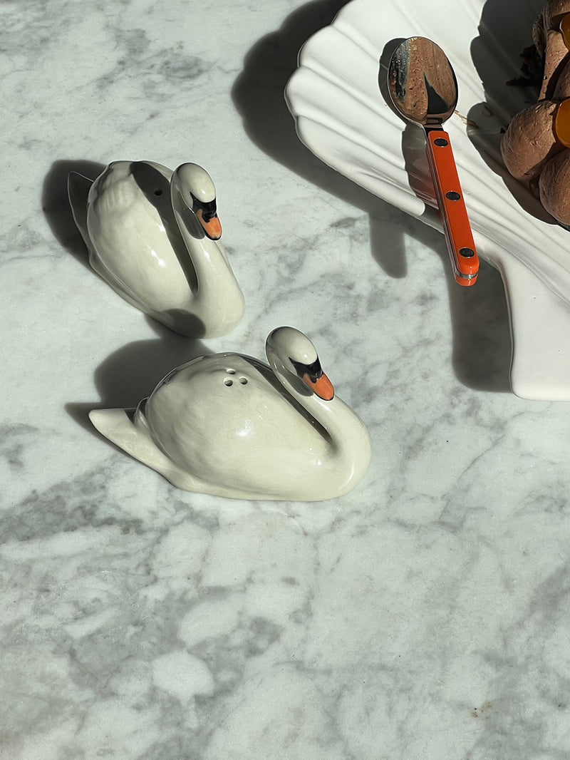 Two white swan shakers with orange beaks and black eye details, placed on a white marble surface next to a white scalloped dish holding a spoon with an orange handle.
