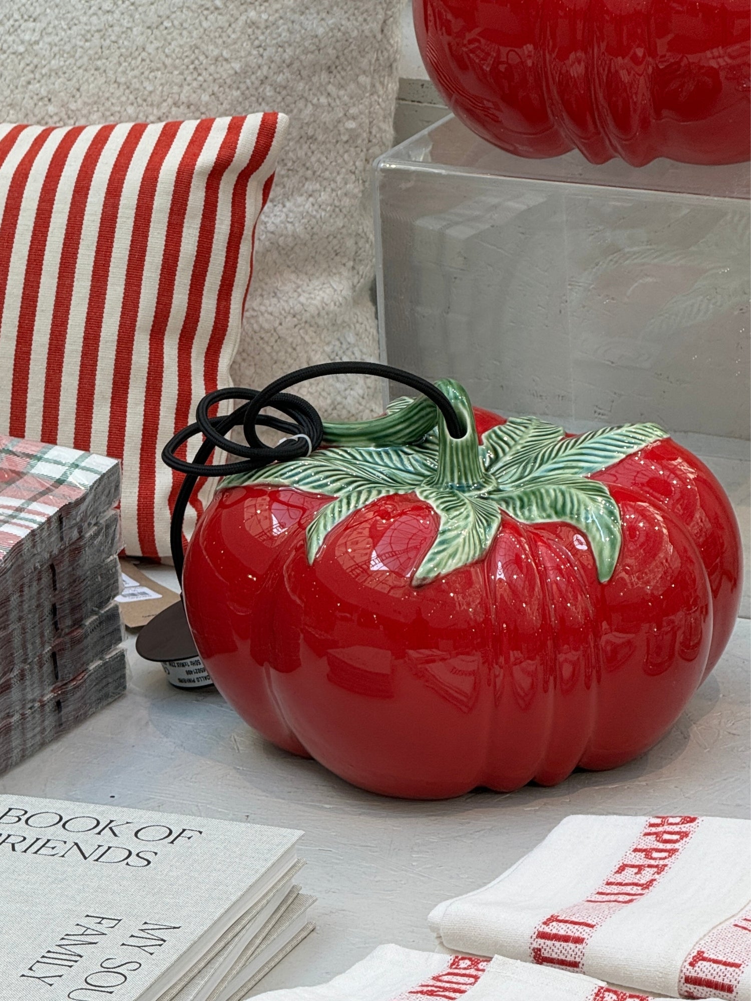 A glossy red ceramic tomato-shaped pendant lamp with a green leafy top and a black loop handle, displayed on a tabletop with a striped pillow, folded white towels with red text, and printed cards in the background.