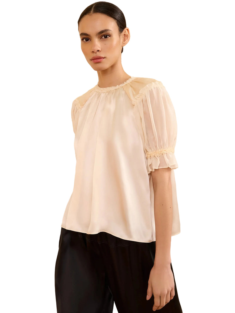 Woman wearing a white blouse with sheer sleeves against a neutral wall.