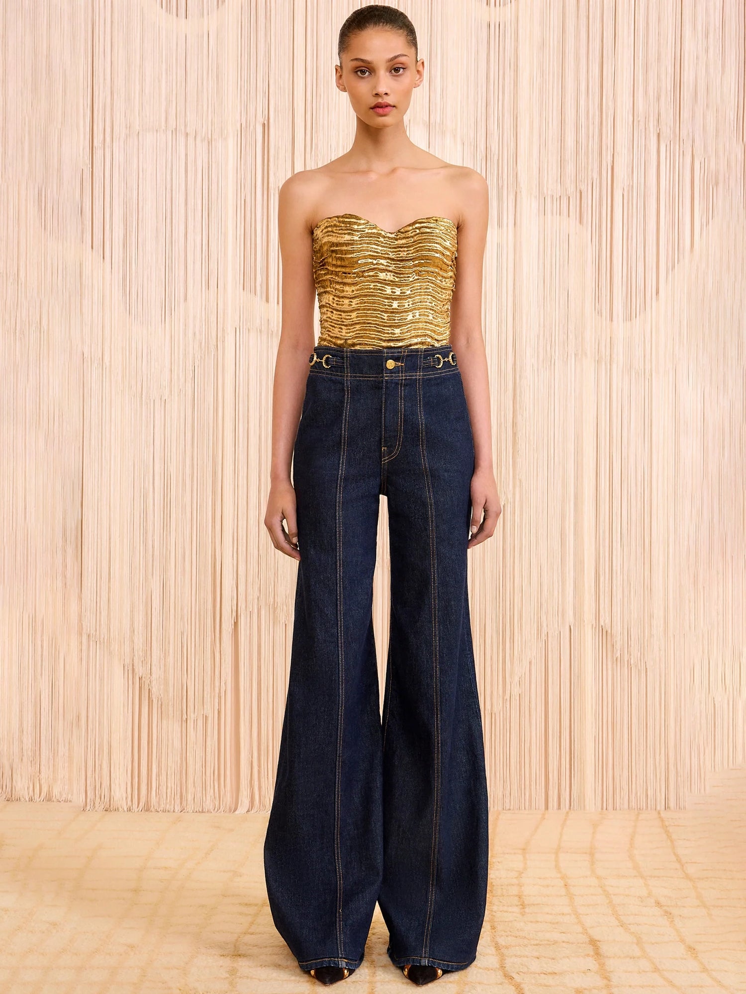 Model wearing a gold strapless top and dark blue jeans against a wooden paneled background