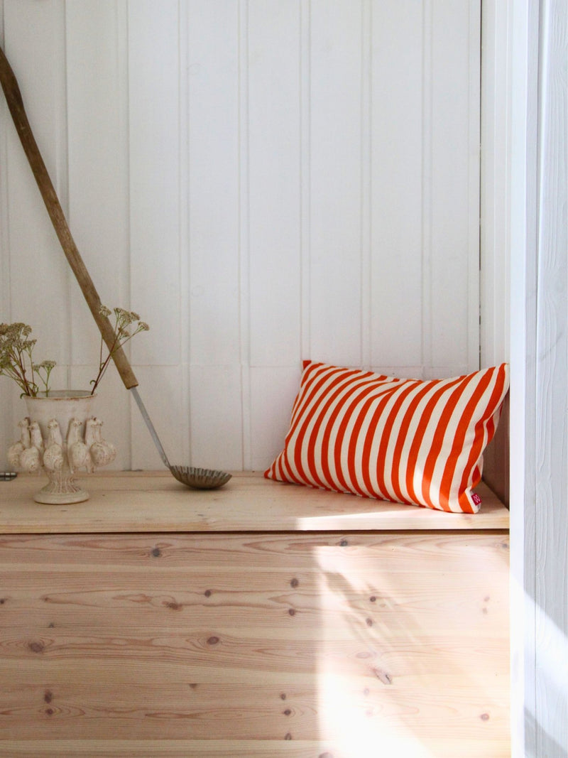An orange and white striped throw pillow resting on a light wood bench against a white paneled wall.