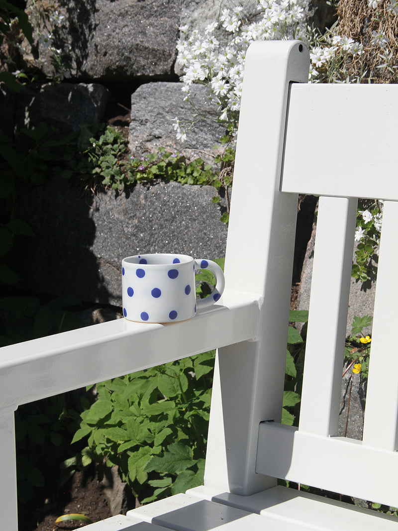 White mug with blue polka dots on a white chair in a garden setting